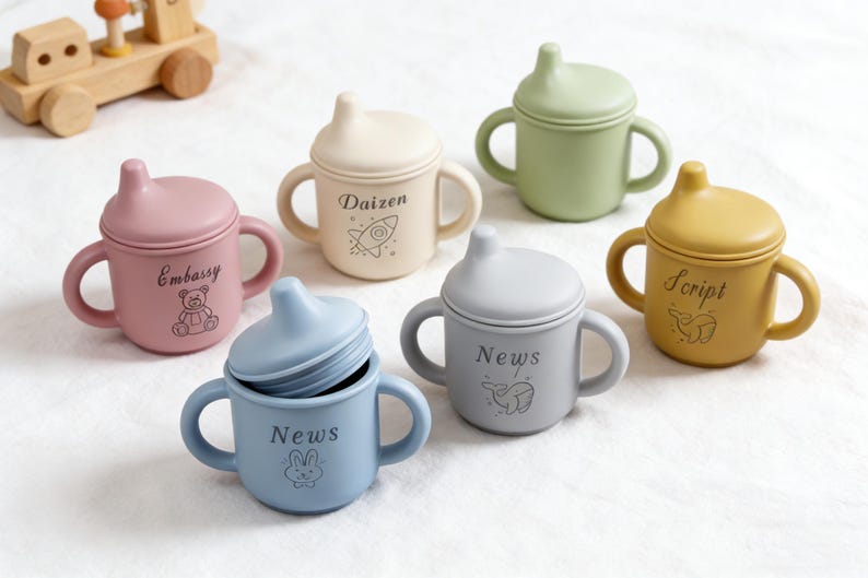 Puede incluir: Colecci&oacute;n de tazas de beb&eacute; de silicona coloridas con asas y tapas. Las tazas son en tonos rosa, crema, verde, amarillo y azul. Cada taza tiene una palabra y un gr&aacute;fico diferentes impresos, como "Embassy" y un osito de peluche.