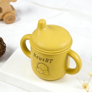 Puede incluir: Vaso para beb&eacute; de color amarillo mostaza con asas y tapa. La taza tiene el texto "NewsBT" y un gr&aacute;fico de sol y nubes. Un coche de juguete de madera y una bola decorativa est&aacute;n en el fondo.
