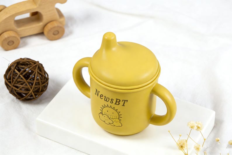 Puede incluir: Vaso para beb&eacute; de color amarillo mostaza con asas y tapa. La taza tiene el texto "NewsBT" y un gr&aacute;fico de sol y nubes. Un coche de juguete de madera y una bola decorativa est&aacute;n en el fondo.