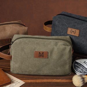 May include: Three rectangular canvas toiletry bags in olive green, charcoal gray, and brown. Each bag features a top zipper, a side handle, and a rectangular leather patch with personalized initials or names. A shaving soap tin and brush are nearby.