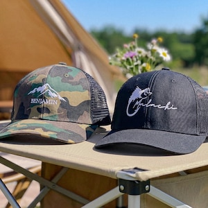 May include: Two trucker hats with mesh backs. The left hat features a green, brown, and black camouflage pattern with an embroidered white mountain graphic and the name Benjamin. The right hat is solid black with a white embroidered fish hook graphic and the name Malachi.