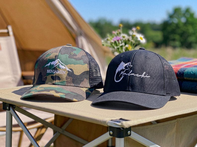 May include: Two trucker hats with mesh backs. The left hat features a green, brown, and black camouflage pattern with an embroidered white mountain graphic and the name Benjamin. The right hat is solid black with a white embroidered fish hook graphic and the name Malachi.