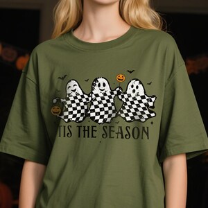 Puede incluir: Camiseta verde oliva con un diseño de Halloween. Presenta tres fantasmas con un patrón de cuadros blancos y negros, sosteniendo calabazas y un globo. El texto "TIS THE SEASON" está impreso debajo de los fantasmas.