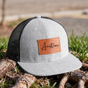 May include: A grey and black trucker cap with a curved peak. The cap has a brown leather patch with the name "Austin" in black script. The cap is on a tree root in a grassy area.