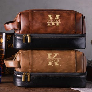 May include: Two personalized leather toiletry bags, one brown and one tan, stacked on top of each other. Each bag has a black base and gold zippers. The top bag is engraved with the name "Michael", and the bottom bag is engraved with the name "Kevin".