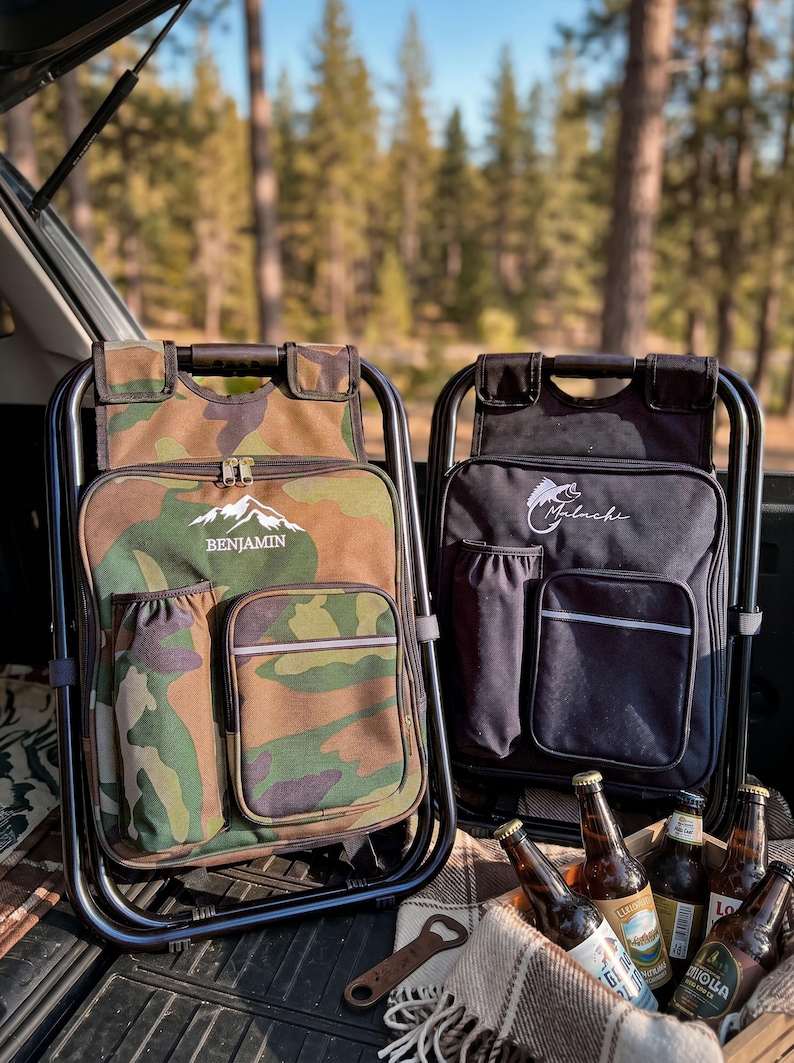 May include: Two folding chair backpacks with black metal frames. One is camouflage print with the text BENJAMIN and a mountain graphic. The other is solid black with a fish graphic and the text Malachi. Both feature side pockets and front pouches.