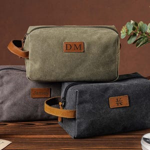 May include: Three rectangular toiletry bags in olive green, grey, and charcoal grey canvas. Each bag features a side handle, a top zipper, and a brown leather patch on the front. The patches are embossed with the letters DM, the name James, and the letters KV.