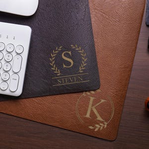 Op de afbeelding: Twee rechthoekige bureauonderleggers met een gestructureerde lederachtige afwerking, een donkerbruine en een lichtbruine. De donkerbruine mat heeft een gouden lauwerkrans-embleem met de letter S en de naam STEVEN. De lichtbruine mat toont een gouden lauwerkrans met de letter K.