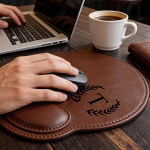 Op de afbeelding: Een bruine leren muismat met polssteun, een zwarte computermuis en de letter "T" in een laurierkransontwerp. Een witte koffiemok, een laptop en een bruin leren notitieboek liggen op het houten bureau.