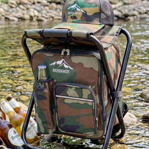 May include: A camouflage print backpack chair with a black metal frame, featuring a matching camouflage baseball cap on top. Both items display a white mountain graphic and the text BENJAMIN. The backpack includes a side pocket holding a glass bottle.