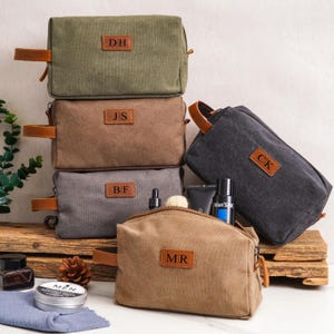 May include: A collection of rectangular canvas toiletry bags in olive green, brown, gray, and tan. Each bag features a brown leather side handle and a central leather patch embossed with two-letter initials: DH, JS, BF, CK, and MR.