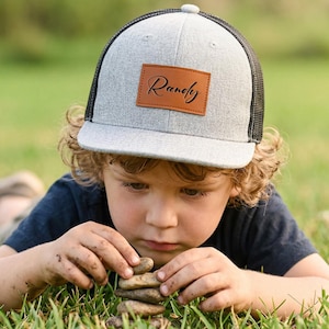 Op de afbeelding: Grijze en zwarte truckerpet met een bruin leren patch met de naam "Randy" in cursief. De pet wordt gedragen door een kind in het gras.