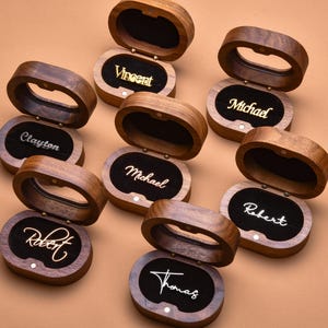 May include: A collection of eight oval-shaped wooden ring boxes with hinged lids. Each box features a dark wood exterior and a black velvet interior lining. The names Vincent, Michael, Clayton, Robert, and Thomas are displayed in gold or white script.