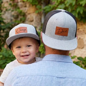 May include: Two grey and black trucker caps. One cap has the name "Cary" on a brown leather patch, and the other cap has the word "Dad" on a similar patch. The caps are worn by a child and an adult.