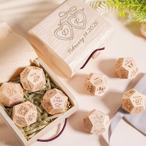 Può includere: Set di dadi incisi in una scatola di legno. Il coperchio della scatola presenta due medaglioni a forma di cuore con le lettere "R" e "D" e la data "February 14, 2026". I dadi sono incisi con suggerimenti di attività.