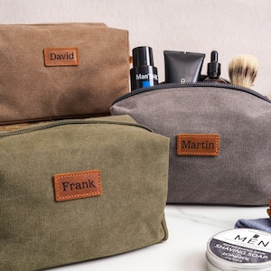 May include: Three canvas toiletry bags in olive green, charcoal gray, and brown. Each bag features a rectangular leather patch with a name embossed: Frank, Martin, and David. The gray bag contains grooming items, and a shaving soap tin is visible.