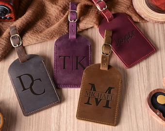 May include: Four leather luggage tags in shades of brown, purple, and burgundy, each with a buckle strap. The tags are embossed with initials and names. The tags are on a wooden surface.