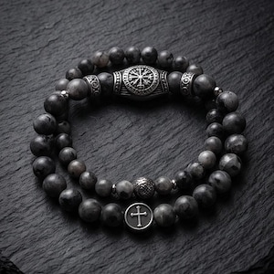 Könnte beinhalten: Ein zweireihiges Armband aus dunkelgrauen, marmorierten Steinperlen. Das obere Band enthält ein silberfarbenes Metallelement mit einem kreisförmigen Vegvisir-Symbol und zwei zylindrische Zwischenstücke. Das untere Band zeigt eine runde silberfarbene Perle mit einem Kreuzsymbol.