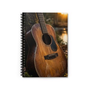 May include: A spiral-bound notebook featuring a cover image of a weathered brown acoustic guitar leaning against a tree trunk. The guitar has a dark pickguard, visible wood grain, and metal strings, with a blurred outdoor sunset background.