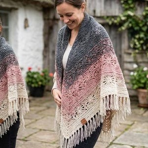 May include: A triangular crochet shawl featuring a gradient color pattern transitioning from dark charcoal gray at the top to dusty rose pink in the middle and cream at the bottom, finished with a long fringe and a small brown leather brand tag.
