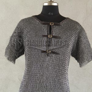 May include: A silver chainmail shirt with short sleeves, displayed on a black mannequin. The shirt features a round neckline and is fastened with three leather straps and brass buckles. The garment is designed for historical reenactment or costume use.