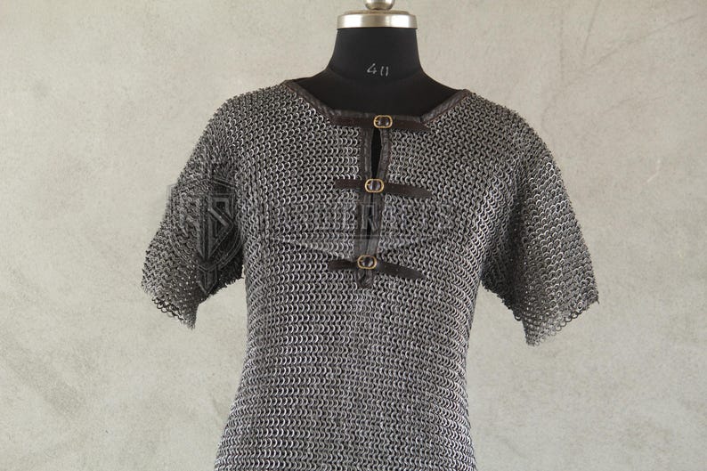 May include: A silver chainmail shirt with short sleeves, displayed on a black mannequin. The shirt features a round neckline and is fastened with three leather straps and brass buckles. The garment is designed for historical reenactment or costume use.