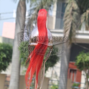 May include: A silver-colored helmet with a long, flowing red plume. The helmet has a reflective surface and is set against a blurred outdoor background. The text "HANDICRAFTS" is visible on the helmet.