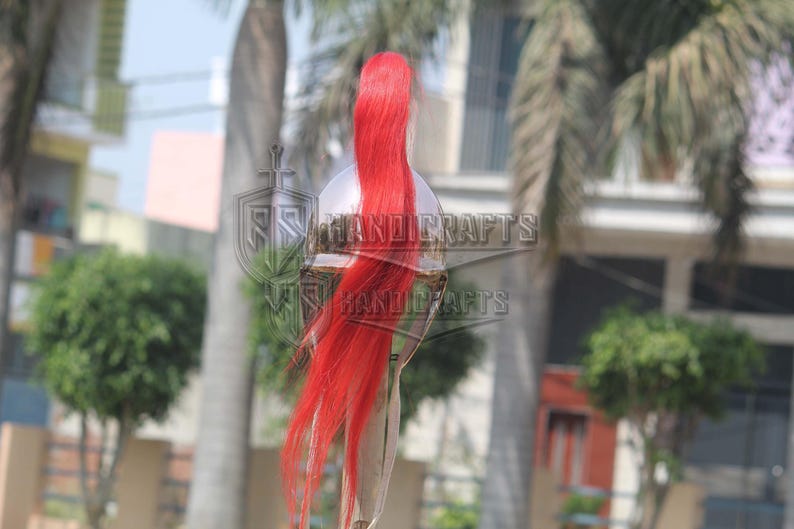 May include: A silver-colored helmet with a long, flowing red plume. The helmet has a reflective surface and is set against a blurred outdoor background. The text "HANDICRAFTS" is visible on the helmet.