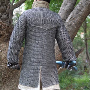 May include: A silver chainmail coat with long sleeves and a split hem. The coat has a high collar and is worn over a light-colored garment. The image is taken outdoors with trees in the background.