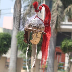 May include: A polished metal Roman helmet with a red plume. The helmet has a curved brow, cheek guards, and a decorative crest. The helmet is mounted on a wooden pole.