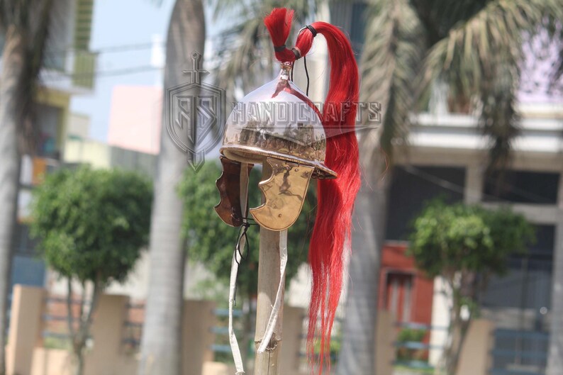May include: A polished metal Roman helmet with a red plume. The helmet has a curved brow, cheek guards, and a decorative crest. The helmet is mounted on a wooden pole.