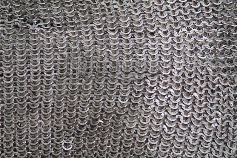 May include: Close-up of a section of chainmail armor. The metal rings are interlocked in a dense pattern, creating a flexible, protective fabric. The silver-colored rings have a slightly weathered appearance.