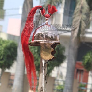 May include: A Roman-style helmet with a polished metal finish and a brass cheek guard. The helmet features a clear visor and is adorned with a long, flowing red plume. The image is set against a blurred outdoor background.