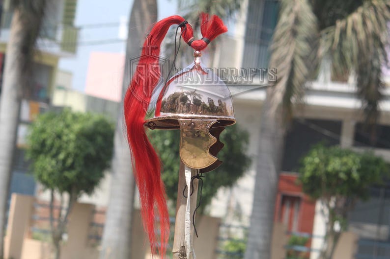 May include: A Roman-style helmet with a polished metal finish and a brass cheek guard. The helmet features a clear visor and is adorned with a long, flowing red plume. The image is set against a blurred outdoor background.