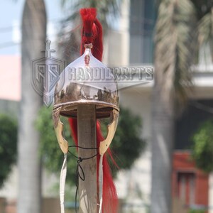 May include: A polished metal helmet with a red plume, likely a historical or costume piece. The helmet has a rounded top and gold-colored cheek guards. The image includes the text "HANDICRAFTS".