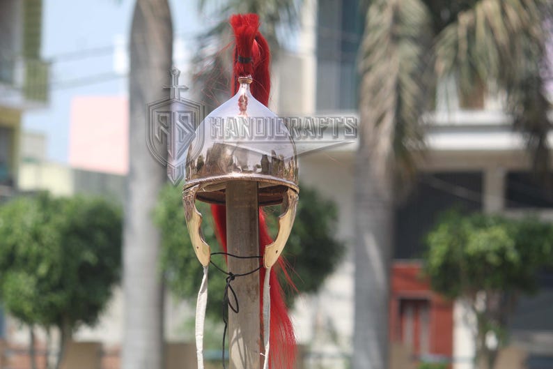 May include: A polished metal helmet with a red plume, likely a historical or costume piece. The helmet has a rounded top and gold-colored cheek guards. The image includes the text "HANDICRAFTS".