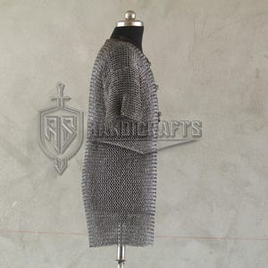 May include: A silver chainmail shirt displayed on a mannequin. The shirt is made of small, interlocking metal rings, creating a flexible, protective garment. The background features a logo and the word "HANDICRAFTS".