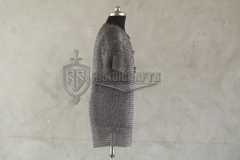 May include: A silver chainmail shirt displayed on a mannequin. The shirt is made of small, interlocking metal rings, creating a flexible, protective garment. The background features a logo and the word "HANDICRAFTS".