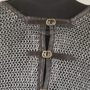 May include: Close-up of a chainmail shirt with a leather collar and straps. The metal chainmail is silver, and the leather straps are brown with gold-colored buckles. The shirt is displayed on a black mannequin.