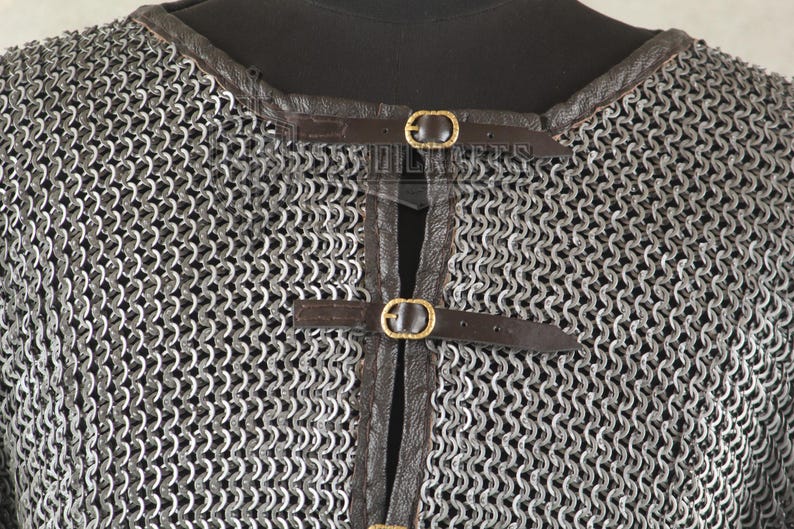 May include: Close-up of a chainmail shirt with a leather collar and straps. The metal chainmail is silver, and the leather straps are brown with gold-colored buckles. The shirt is displayed on a black mannequin.