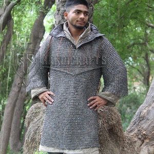 May include: A silver chainmail shirt with a hood and long sleeves. The shirt has a tunic-like design and is worn over a light-colored garment. The image is taken outdoors with a background of trees.
