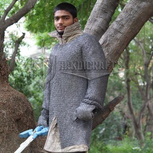 May include: A person wearing a chainmail shirt with a high collar and long sleeves. The chainmail is a silver color. The person is holding a blue and white sword. The background is a natural outdoor setting with trees.
