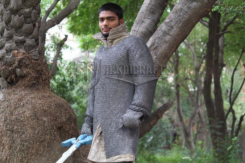May include: A person wearing a chainmail shirt with a high collar and long sleeves. The chainmail is a silver color. The person is holding a blue and white sword. The background is a natural outdoor setting with trees.