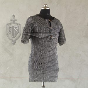 May include: A short-sleeved chainmail shirt displayed on a mannequin. The shirt is made of small, interlocking metal rings, creating a flexible, protective garment. It features a round neckline and leather straps with buckles. The background is a neutral gray.