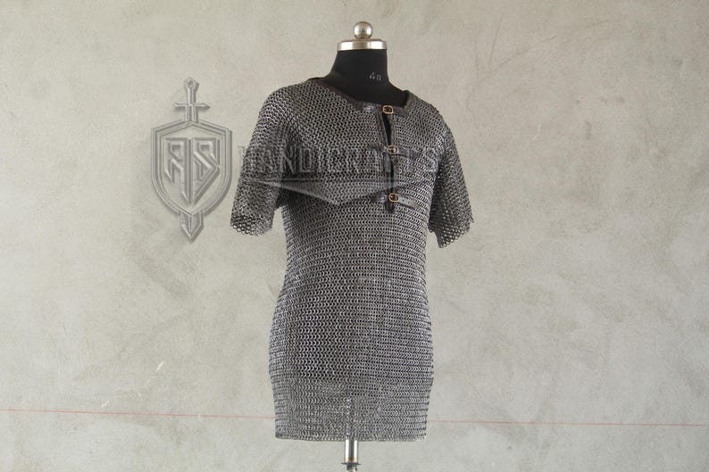 May include: A short-sleeved chainmail shirt displayed on a mannequin. The shirt is made of small, interlocking metal rings, creating a flexible, protective garment. It features a round neckline and leather straps with buckles. The background is a neutral gray.