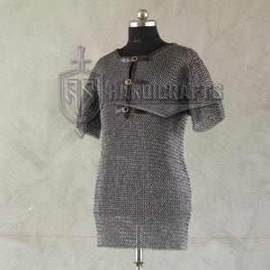 May include: A silver chainmail shirt with short sleeves, displayed on a black mannequin. The shirt has a round neckline and is fastened with leather straps. The metal links create a textured surface, and the garment is designed for historical reenactment or costume use.