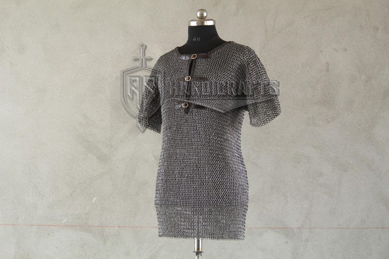 May include: A silver chainmail shirt with short sleeves, displayed on a black mannequin. The shirt has a round neckline and is fastened with leather straps. The metal links create a textured surface, and the garment is designed for historical reenactment or costume use.