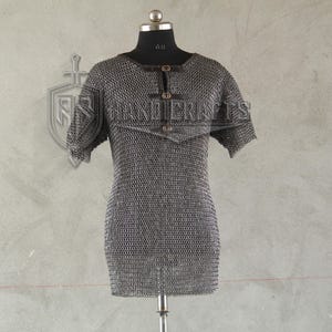 May include: A silver chainmail shirt with short sleeves, displayed on a mannequin. The shirt has a round neckline and is fastened with three small, dark-colored buttons. The background features a shield emblem.