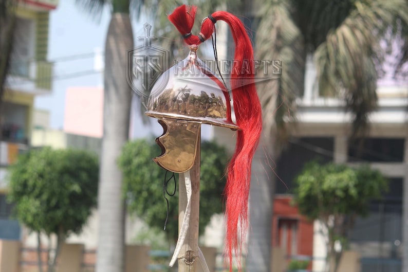 May include: A polished metal helmet with a red plume. The helmet has a curved design with a brass-colored cheek guard. The plume is long and flowing, adding a striking visual element. The helmet is mounted on a wooden stand.
