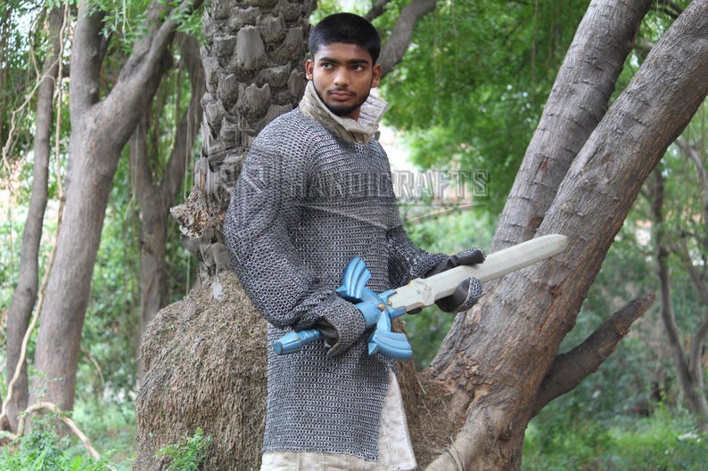 May include: A person wearing a chainmail shirt and holding a wooden sword with a blue hilt. The sword has a light-colored blade. The image is set outdoors with trees in the background. The text "HANDICRAFTS" is visible.
